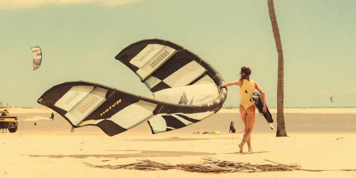 Kitesurfista atravessando a praia entre as asas de kite e os coqueiros